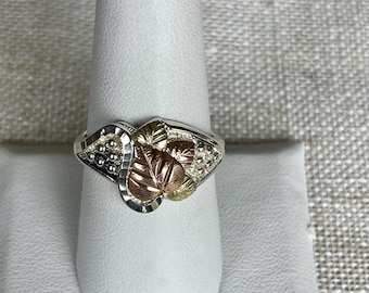 Puede incluir: Un anillo de plata con un diseño de hojas de oro y cobre. El anillo está en un expositor blanco.