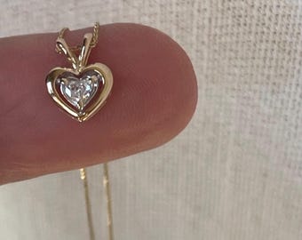 May include: A gold heart-shaped pendant necklace featuring a single clear gemstone set in the center. The pendant is suspended from a delicate gold chain, showcasing a polished metal frame that surrounds the central stone in a classic design.