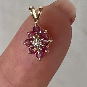 Puede incluir: Un colgante de oro con un diamante blanco redondo central rodeado por un grupo de ocho rubíes de color rojo intenso dispuestos en un patrón floral. El colgante incluye una anilla de oro en la parte superior para sujetarlo a una cadena.