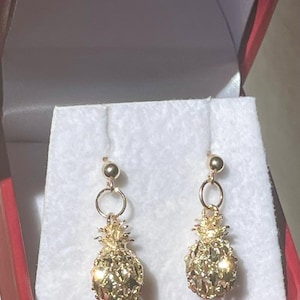 Könnte beinhalten: Ein Paar goldfarbene Ohrhänger mit einem texturierten, dreidimensionalen Ananas-Anhänger, der an einem kleinen kreisförmigen Ring und einem Kugelstecker befestigt ist. Die Ohrringe sind auf einem weißen Stoffeinsatz in einer roten Schachtel.