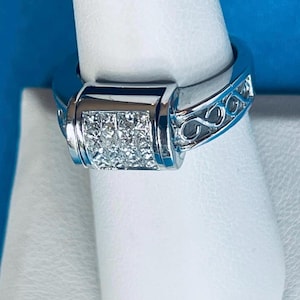 May include: A silver-toned ring featuring a square-shaped centerpiece embedded with multiple small clear stones. The band displays an intricate openwork scroll pattern along the sides, providing a detailed texture to the metallic surface.