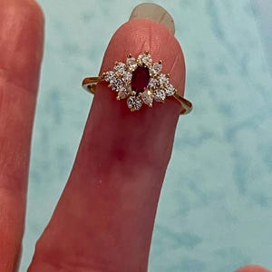 Puede incluir: Un anillo de oro con una piedra roja central de corte ovalado rodeada por un grupo de doce pequeñas piedras blancas claras y redondas dispuestas en un patrón floral. La banda es delgada y de oro pulido, creando un aspecto delicado y refinado.
