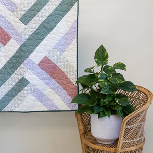 May include: A handmade quilt with a geometric design in shades of white, green, lavender, and mauve, hanging from a copper rod. The quilt is displayed with a wicker chair and potted plants, creating a cozy, inviting atmosphere.