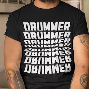Op de afbeelding: Een zwart T-shirt met een wit grafisch ontwerp waarop meerdere keren "DRUMMER" staat in een herhalend patroon, waarbij elk woord iets verschoven is ten opzichte van het woord erboven.