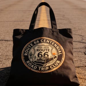 May include: Black tote bag with a Route 66 Centennial Road Trip Club design. The design features a vintage car, cactus, and the years 1926-2026. The bag is sitting on a road.