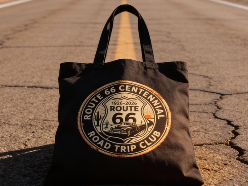 May include: Black tote bag with a Route 66 Centennial Road Trip Club design. The design features a vintage car, cactus, and the years 1926-2026. The bag is sitting on a road.