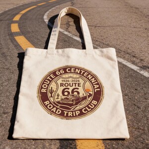 May include: Cream-colored tote bag featuring a Route 66 Centennial Road Trip Club design. The circular graphic includes a vintage car, cactus, and the years 1926-2026. The bag is sitting on a road.