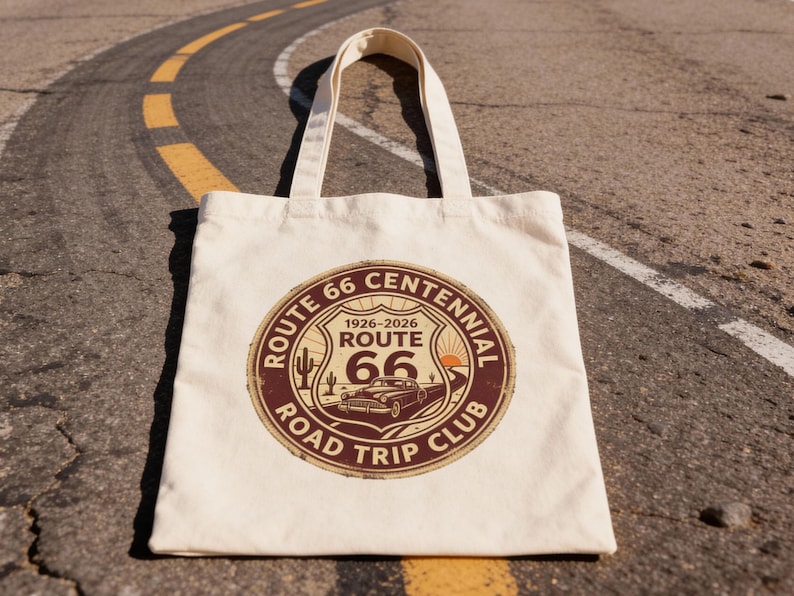 May include: Cream-colored tote bag featuring a Route 66 Centennial Road Trip Club design. The circular graphic includes a vintage car, cactus, and the years 1926-2026. The bag is sitting on a road.