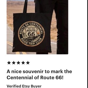 May include: Black tote bag featuring a Route 66 Centennial design. The circular emblem includes the text "Route 66 Road Trip Club" and the years "1926-2026". The design is in shades of brown, white, and black. The bag is held by a person.