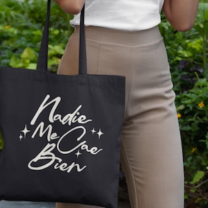 May include: Black tote bag with the Spanish phrase "Nadie Me Cae Bien" in white script. The bag has long black handles and is held by a person wearing a white top and tan pants. The background features green foliage.