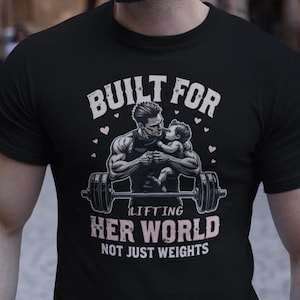 May include: Black t-shirt with a graphic of a muscular man lifting weights with a baby in his arms. The text "Built for lifting her world, not just weights" is printed on the shirt.