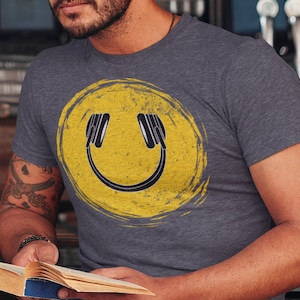 May include: A gray t-shirt with a yellow smiley face graphic. The smiley face has headphones on and a distressed, vintage look.