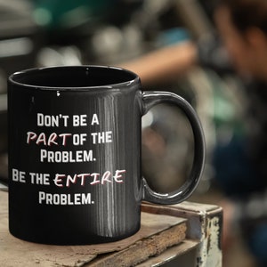 Op de afbeelding: Zwarte keramische mok met de tekst "DON'T BE A PART OF THE PROBLEM. BE THE ENTIRE PROBLEM." in witte en rode letters. De mok heeft een groot handvat en staat op een houten oppervlak.
