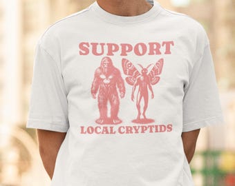 May include: White t-shirt with a pink graphic design that says "SUPPORT LOCAL CRYPTIDS". The graphic features a Bigfoot and a mothman.