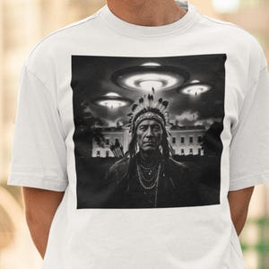 May include: A black and white graphic t-shirt featuring a Native American man wearing a headdress, with flying saucers in the background. The image is set against a white house.
