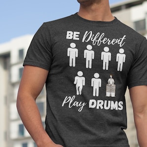 Könnte beinhalten: Ein dunkelgraues T-Shirt mit dem Text "BE DIFFERENT" und einer Reihe von Strichmännchen. In der unteren Reihe spielt ein Strichmännchen Schlagzeug mit dem Text "Play DRUMS".