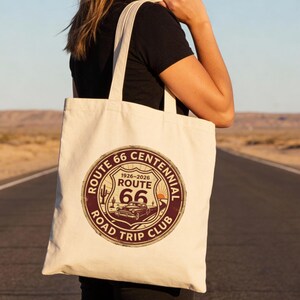 May include: Cream-colored Route 66 Centennial Road Trip Club tote bag. The circular graphic features a vintage car, cactus, and the text "Route 66" and "1926-2026". The bag is carried on a road with a desert backdrop.