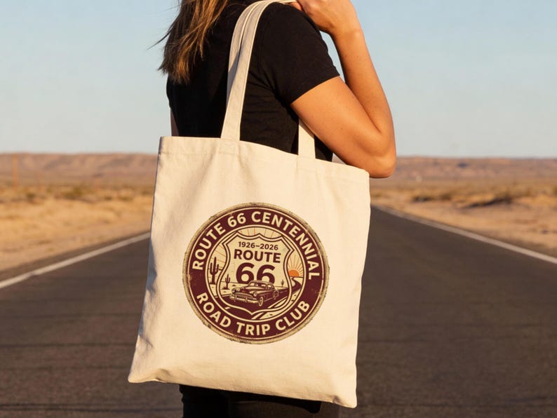 May include: Cream-colored Route 66 Centennial Road Trip Club tote bag. The circular graphic features a vintage car, cactus, and the text "Route 66" and "1926-2026". The bag is carried on a road with a desert backdrop.