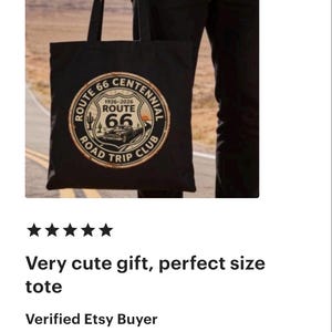 May include: Black tote bag featuring a Route 66 Centennial design with a classic car illustration. The design includes the text "Route 66 Road Trip Club" and the years 1926-2026. The bag is held by a person.