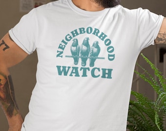 Puede incluir: Camiseta blanca con un gráfico azul verdoso de tres palomas posadas en un alambre con el texto "Neighborhood Watch" debajo.