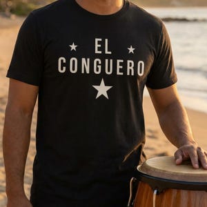 Puede incluir: Camiseta negra con el texto blanco "EL CONGUERO", con estrellas arriba y abajo. La persona está de pie junto a un tambor conga en una playa.