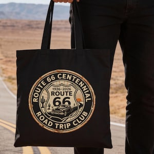 May include: Black tote bag featuring a Route 66 Centennial design. The design includes a vintage car, desert landscape, and the text "Route 66 Road Trip Club" with the years 1926-2026. The bag is held in front of a desert road.
