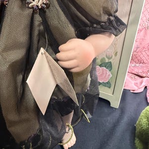 以下が含まれることがあります： 黒と金のドレスを着たヴィンテージドール。装飾的なレースの襟とビーズのアクセントが特徴です。人形の腕が見え、ドレスにはタグが付いています。ドレスは、金色の生地の上にシアーブラックの生地で作られています。