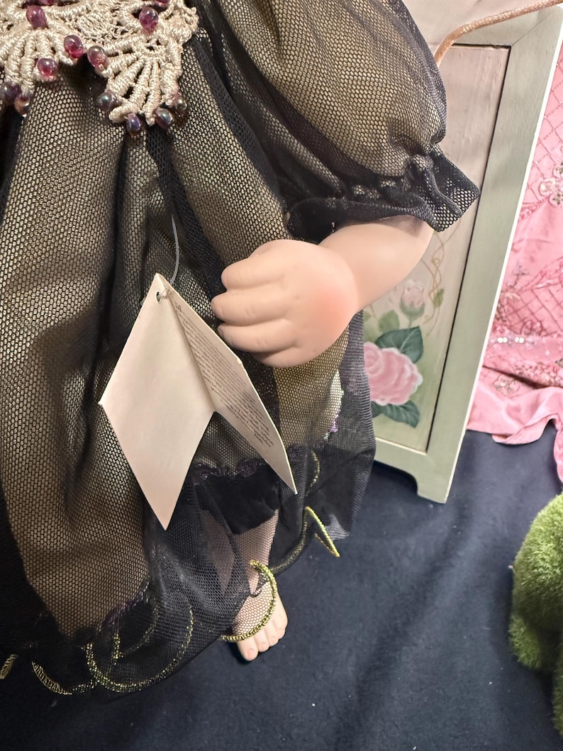 以下が含まれることがあります： 黒と金のドレスを着たヴィンテージドール。装飾的なレースの襟とビーズのアクセントが特徴です。人形の腕が見え、ドレスにはタグが付いています。ドレスは、金色の生地の上にシアーブラックの生地で作られています。