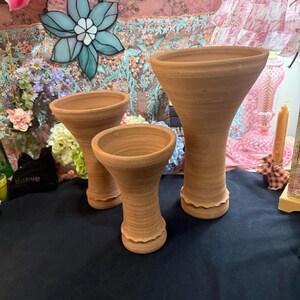May include: Three terracotta vases of varying heights, each featuring a wide, flared opening, a slender neck with horizontal ridged textures, and a decorative scalloped base. The natural clay material has a matte, earthy brown finish.