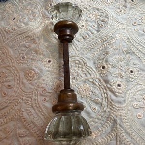 May include: A door knob set featuring two clear, faceted glass knobs with brass-toned metal bases connected by a long, square-shaped metal spindle. The glass knobs have a ribbed, circular design, and the metal components show a weathered, aged finish.