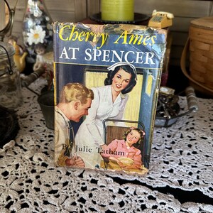 May include: Vintage paperback book titled "Cherry Ames at Spencer" by Julie Tatham. The cover features a nurse, a doctor, and a young girl in a hospital setting.