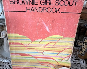 May include: A vintage red and yellow Brownie Girl Scout Handbook with the title "BROWNIE GIRL SCOUT HANDBOOK" printed in black.