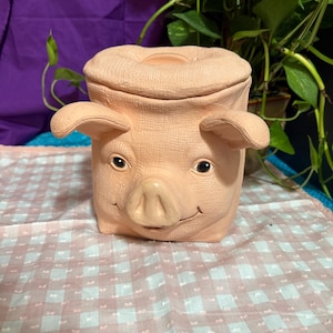 May include: A light peach-colored ceramic canister shaped like a pig's head with a textured surface. The canister features a removable lid, two protruding ears, a snout, and a subtle smiling expression with dark eyes, designed for storage.
