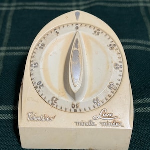 May include: Off-white vintage kitchen timer with a curved top and a central dial. The timer features a silver-colored indicator and a circular dial with numbers. The words "Robertshaw" and "Lux minute minder" are printed on the front.
