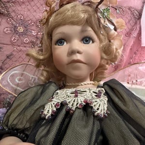 以下が含まれることがあります： ブロンドの髪と青い目の磁器人形。花の髪飾りと繊細な妖精の羽で飾られています。人形は、ビーズの襟が付いた黒と金のドレスを着ています。人形の表情は穏やかです。