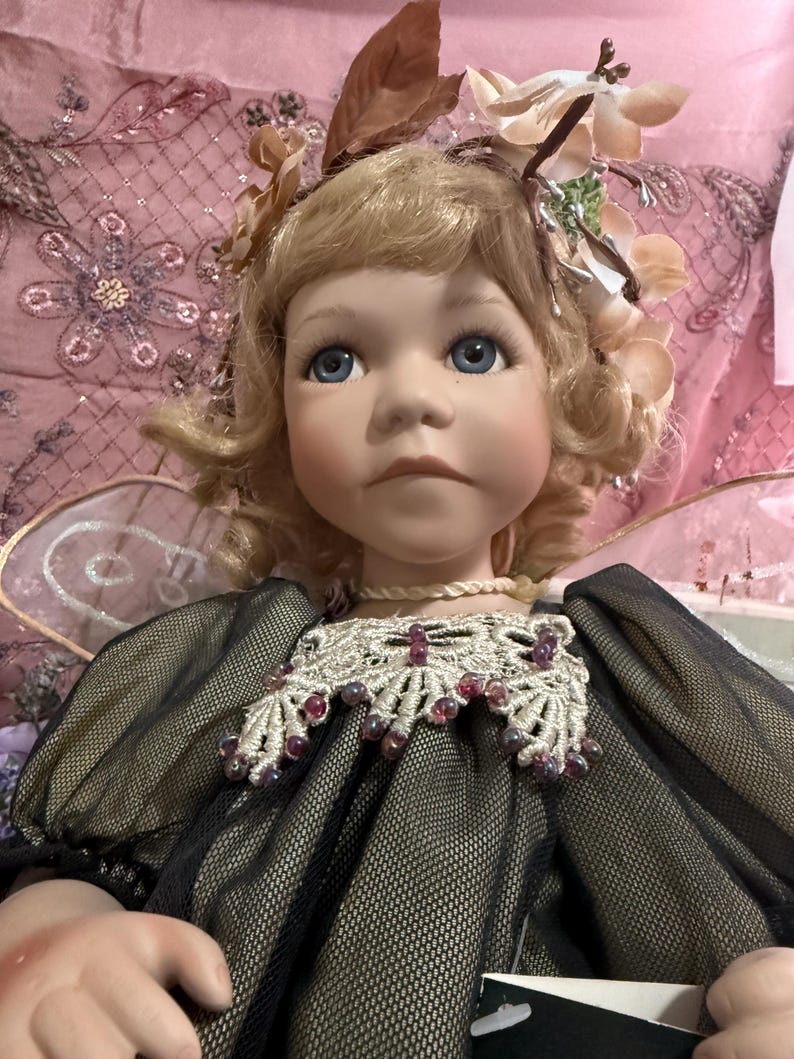 以下が含まれることがあります： ブロンドの髪と青い目の磁器人形。花の髪飾りと繊細な妖精の羽で飾られています。人形は、ビーズの襟が付いた黒と金のドレスを着ています。人形の表情は穏やかです。