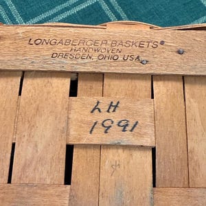以下が含まれることがあります： 「LONGABERGER BASKETS HANDWOVEN DRESDEN, OHIO USA」と「LH 1991」の文字が書かれた、木製の編みかごのクローズアップ。 かごは明るい茶色の木のストリップで作られています。