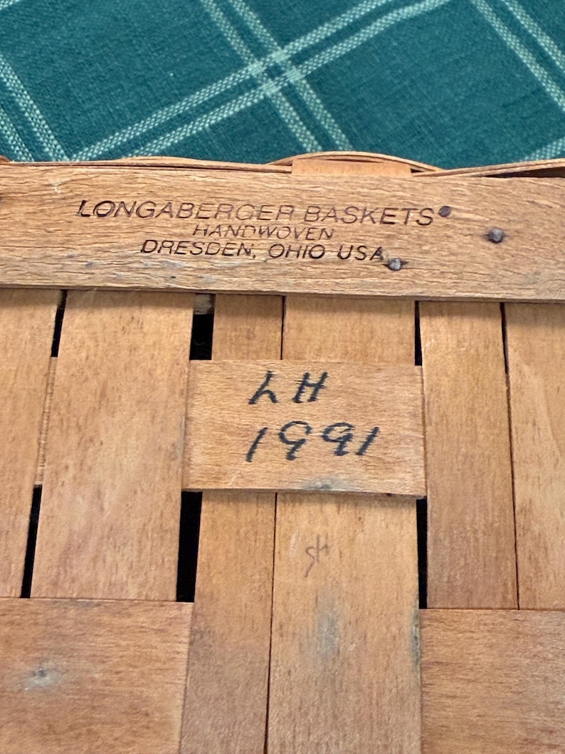 以下が含まれることがあります： 「LONGABERGER BASKETS HANDWOVEN DRESDEN, OHIO USA」と「LH 1991」の文字が書かれた、木製の編みかごのクローズアップ。 かごは明るい茶色の木のストリップで作られています。