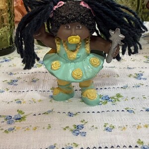 May include: A small doll with dark skin and black yarn hair styled with pink bows. The doll wears a light blue dress with yellow rose accents, a yellow necklace, and yellow shoes. It holds a silver star-shaped wand and has a yellow pacifier.