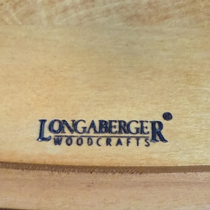 以下が含まれることがあります： 明るい色の木材の表面のクローズアップ。濃い青色で「LONGABERGER WOODCRAFTS」の文字が書かれています。 木目は見え、テキストは中央に配置されています。