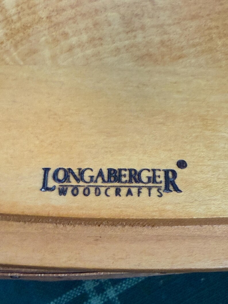 以下が含まれることがあります： 明るい色の木材の表面のクローズアップ。濃い青色で「LONGABERGER WOODCRAFTS」の文字が書かれています。 木目は見え、テキストは中央に配置されています。
