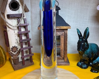 Könnte beinhalten: Eine hohe, zylindrische Solitärvase aus klarem Glas mit einem tiefblauen Innenkern. Die Vase hat eine schräg geschnittene Öffnung und einen kleinen runden rot-weißen Aufkleber nahe der Basis, platziert auf einer runden Holzunterlage.