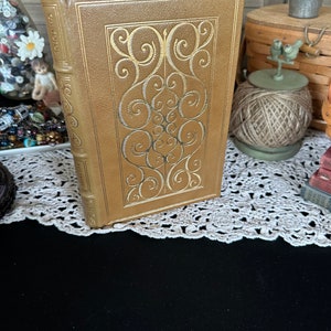 May include: A brown leather-bound book with gold embossed details. The book has a decorative design on the front cover with a swirling pattern.