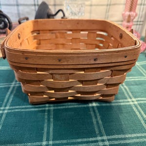 以下が含まれることがあります： 正方形の形をした、長方形の薄茶色の織りバスケット。 バスケットは織り模様と、小さな金属製の鋲で固定された補強された縁が特徴です。 緑と白の格子縞の表面に置かれています。 収納に最適です。