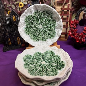 May include: A stack of ceramic plates featuring a raised, textured white cabbage leaf border and a central green cabbage leaf design. The plates have an irregular, wavy edge and a glossy finish, showcasing a detailed, embossed botanical pattern.