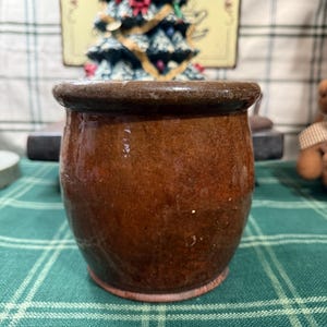 May include: A small, round, brown ceramic pot with a slightly flared rim. The pot has a glossy finish and a reddish-brown base. The background includes a Christmas tree decoration and a green and white plaid tablecloth.