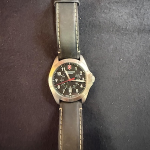 May include: A Victorinox Swiss Army wristwatch featuring a round stainless steel case, a black dial with white Arabic numerals, a red second hand, and a date window at the six o'clock position. The watch includes a black leather strap with light stitching.