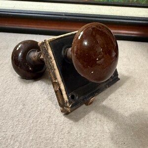 May include: A vintage door knob set featuring two brown ceramic knobs. The knobs are round and have a glossy finish. The set includes a black square plate with a distressed, aged appearance. The hardware is likely made of metal.