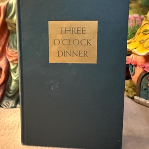 May include: A hardcover book with a dark teal cover and a gold rectangular label. The label reads "THREE O'CLOCK DINNER" in a serif font. The book is standing upright, suggesting it is a vintage cookbook or a book about dining.