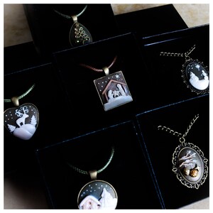 May include: A collection of Christmas-themed pendant necklaces displayed in black boxes. The pendants feature various designs, including a reindeer in a snowy landscape, a nativity scene, and a tree. The necklaces have bronze-colored metal frames and colored cords.